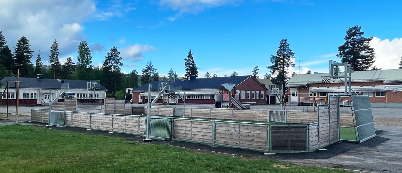 Multiarena av trä med basketstativ, arenamål och konstgräs, placerad på en skolgård.