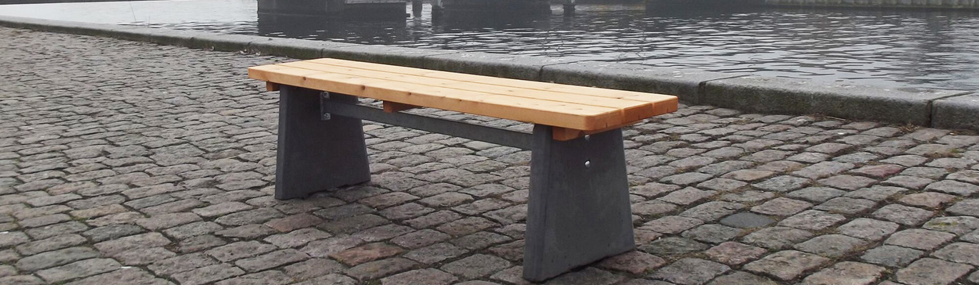 Parkbenk av betong og tre, på kaien med en bro i bakgrunnen.